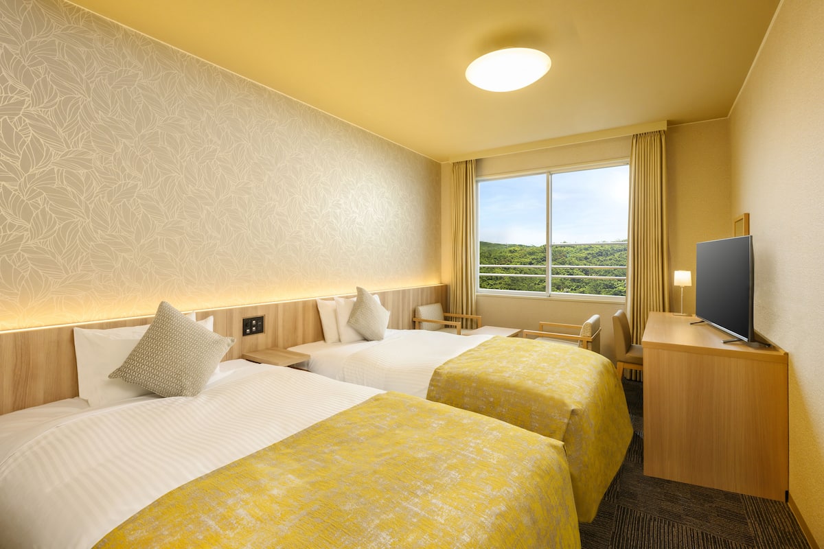 Habitación de hotel con dos camas, un escritorio con televisor y una ventana con vista a áreas verdes.