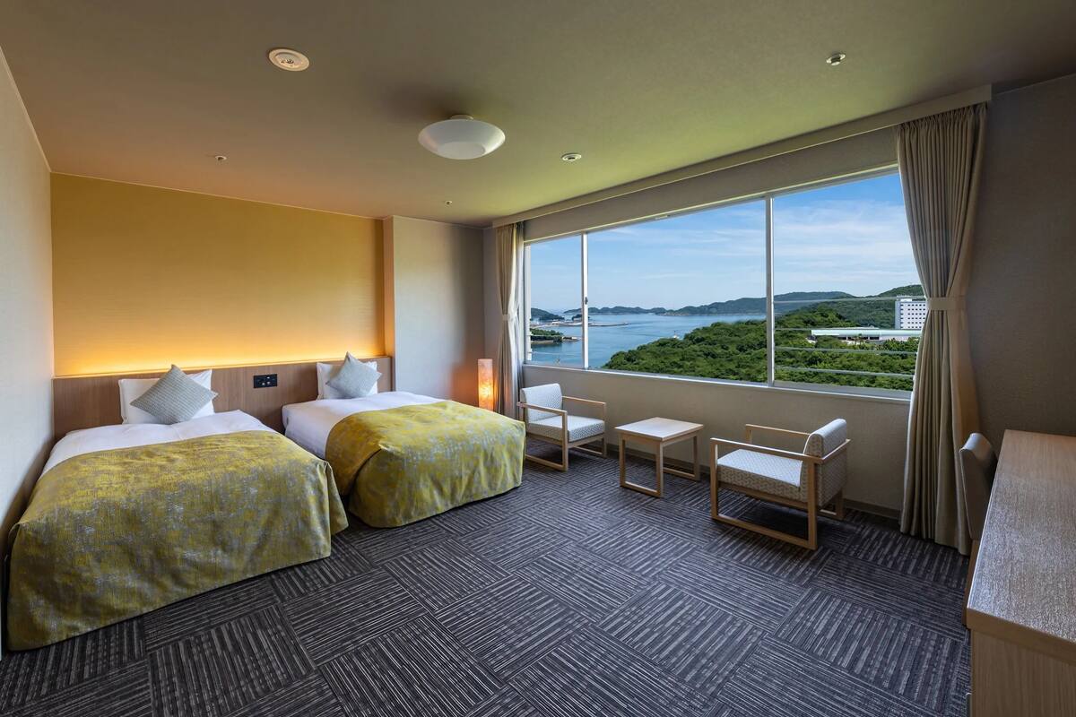 Habitación de hotel con dos camas, una mesita, una silla y vista a un cuerpo de agua y follaje.