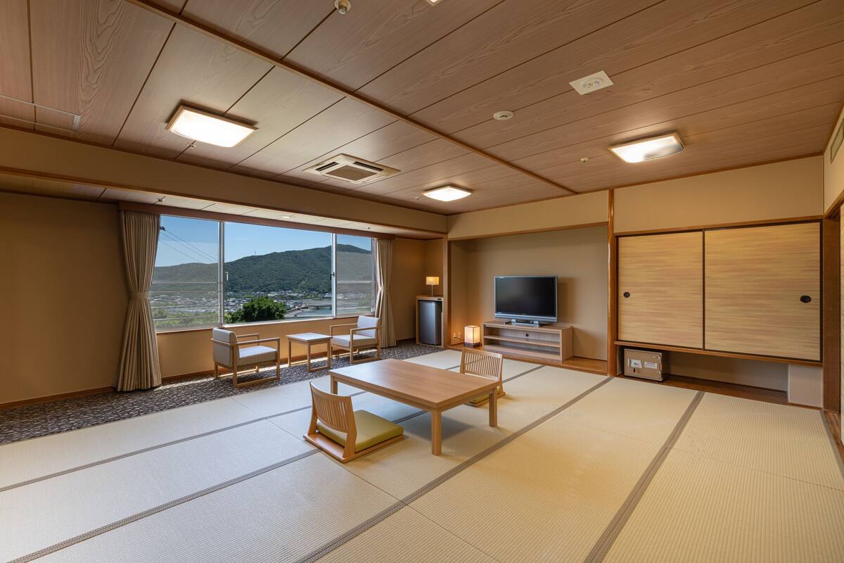 Habitación de hotel con suelo de tatami, televisor, escritorio con sillas, ventana grande con vista a la montaña y un armario.