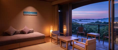 Una habitación moderna con sofá, sillas y una mesa, que cuenta con un amplio ventanal corredero a un balcón con vistas al mar y edificios a la distancia.