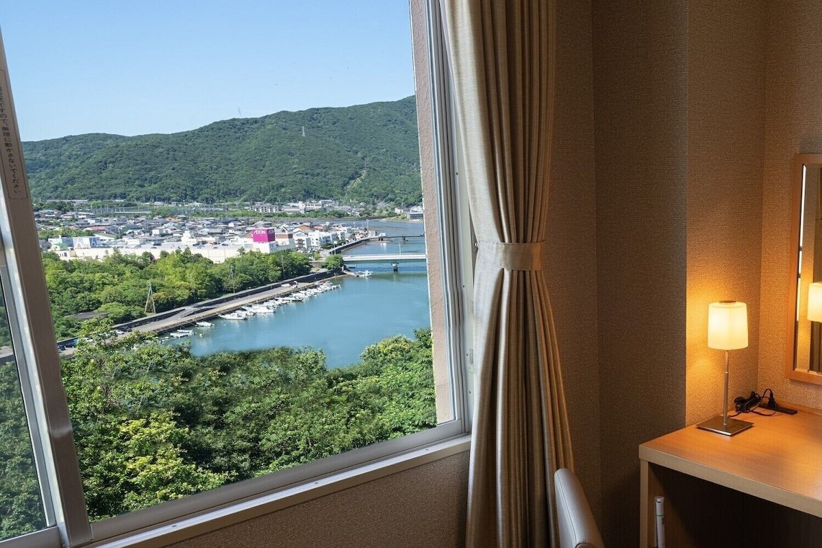 Habitación de hotel con un ventanal que ofrece vistas a un puerto deportivo, barcos y una ciudad al otro lado del río.