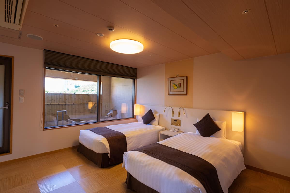Habitación de hotel con dos camas, un ventanal grande y una mesita de noche con lámpara.
