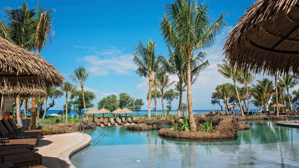 Ein Poolbereich eines Resorts mit Liegestühlen, Palmen und Blick auf den Ozean.