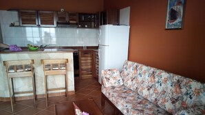 A living room with a sofa, a coffee table with flowers, a kitchen area with cabinets and a refrigerator, and a tiled floor.