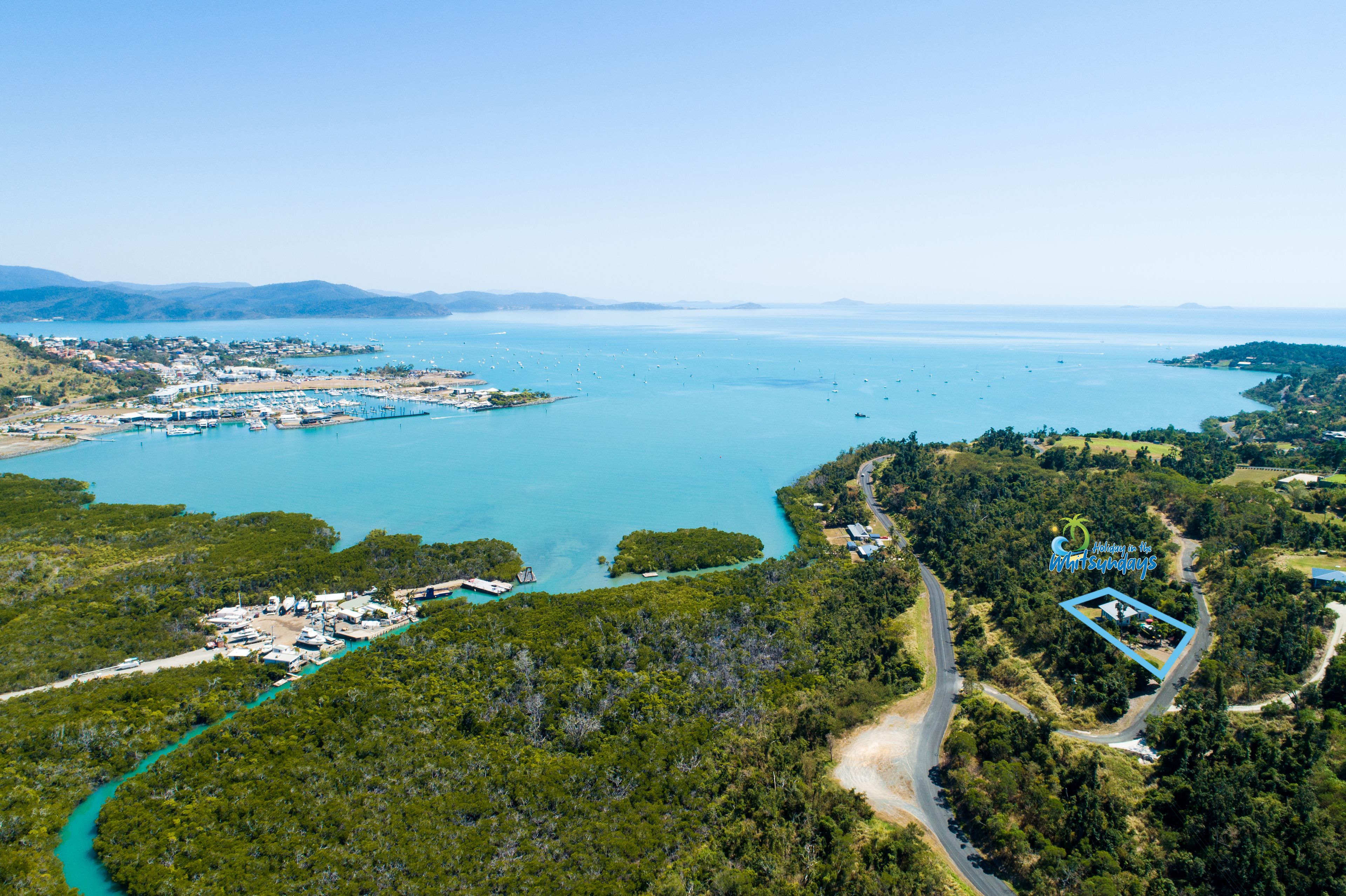 An aerial view of a coastal area with a marina, dense forest, and a clear sky.