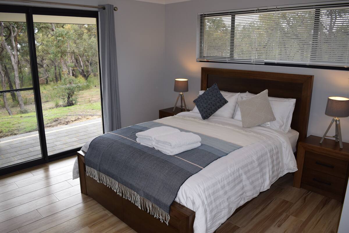 A bedroom with a large window, a bed with a grey and white striped blanket, two bedside lamps, and a view of greenery outside.