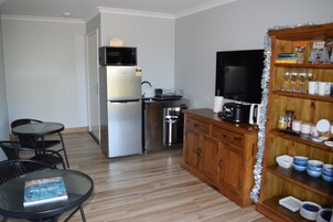 Cocina compacta equipada con refrigerador, microondas, cafetera y televisión. Hay un comedor con mesa y sillas, y una alacena con vajilla y vasos.