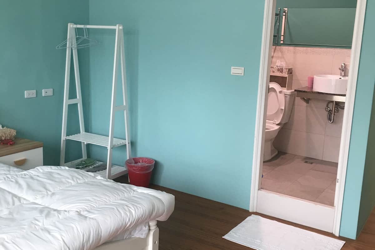 Une chambre avec un lit, un climatiseur blanc, une échelle blanche, une poubelle rouge et une salle de bain visible à travers une porte ouverte.
