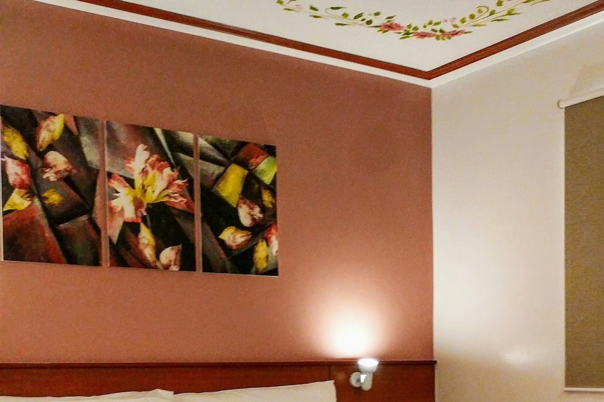 Une chambre avec un lit en bois, une lampe fixée au mur et un tableau représentant des motifs floraux.