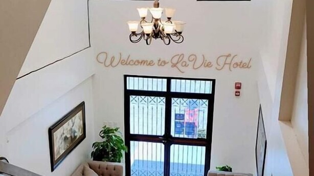 Um hall de hotel com uma escadaria, um candelabro e uma placa com a mensagem 'Welcome to La Vie Hotel'.