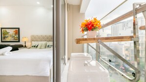 A hotel room with a bed, a window with a view of the city, a small stool, and a vase with flowers.