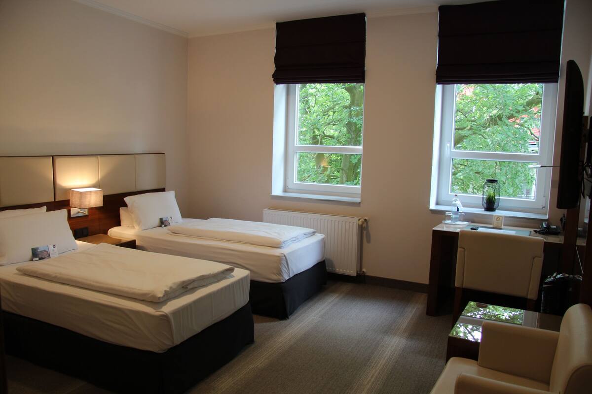 Quarto de hotel com duas camas, uma escrivaninha, uma cadeira e janela com vista para árvores.