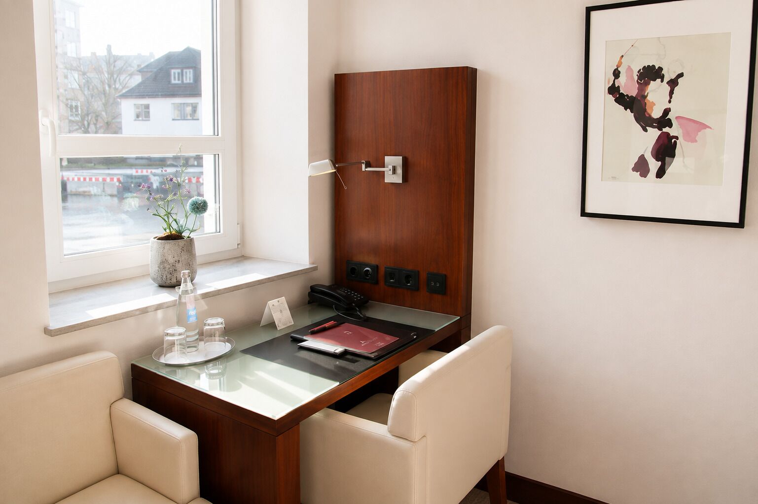 Une chambre d’hôtel avec un bureau en bois, un téléphone, un tableau encadré et une fenêtre donnant sur des immeubles.