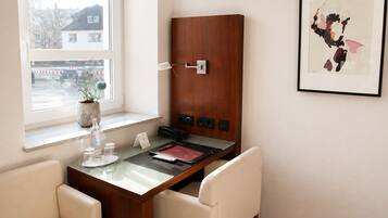 Une chambre d’hôtel avec un bureau en bois, un téléphone, un tableau encadré et une fenêtre donnant sur des immeubles.