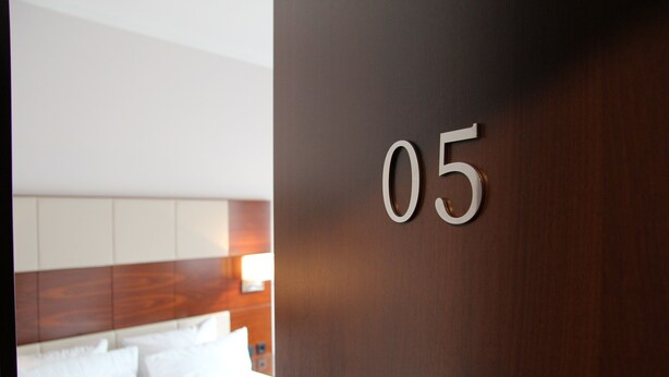 Habitación de hotel con cabecera de madera, cama con ropa blanca y el número '05' colocado en la pared.