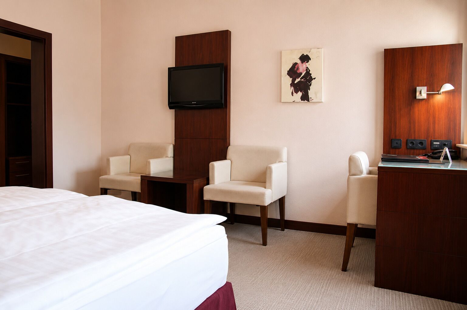 A hotel room with a bed, two armchairs, a desk with a lamp, a television, and a painting on the wall.