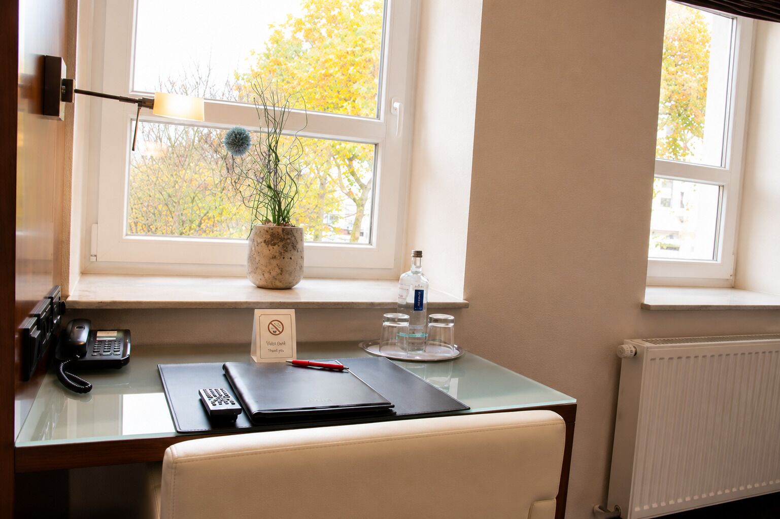 Une chambre d’hôtel équipée d’un bureau, d’un téléphone, d’un ordinateur portable, d’une plante en pot et d’une fenêtre donnant sur des arbres.