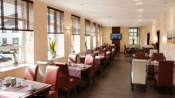 Un comedor de hotel con varias mesas y sillas, ventanas amplias con persianas y un televisor de pantalla plana montado en la pared.