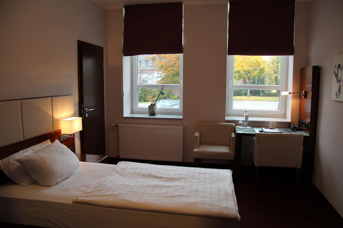 Quarto de hotel com cama, escrivaninha, cadeira, radiador e duas janelas com cortinas.