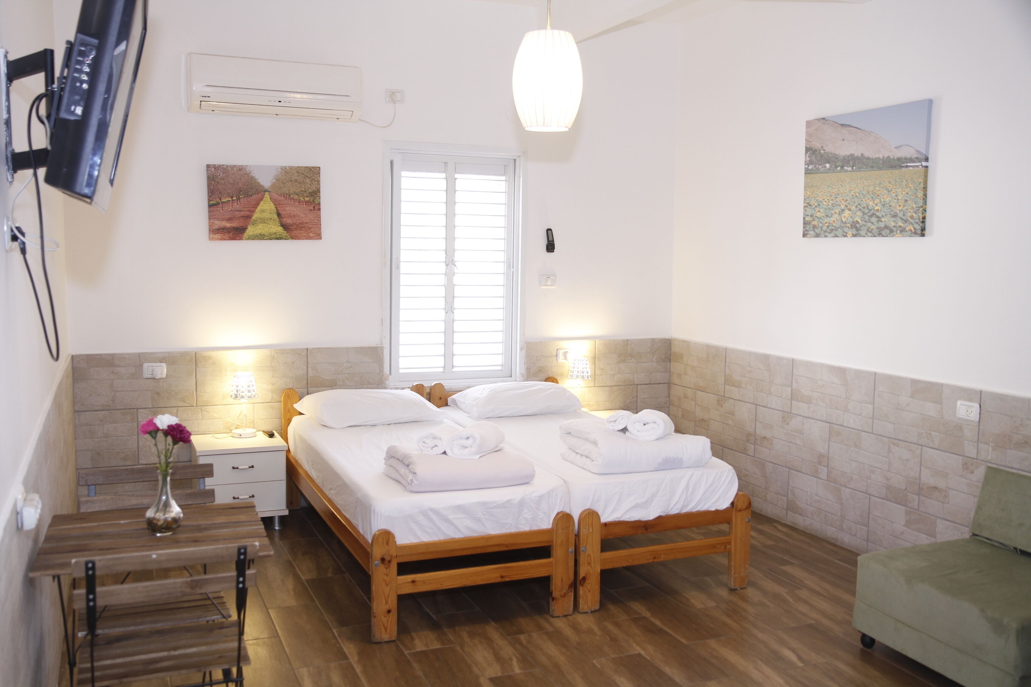Una habitación de hotel con una cama de madera, una mesita de noche, una mesilla con un jarrón de flores, un sillón verde y un cuadro en la pared.