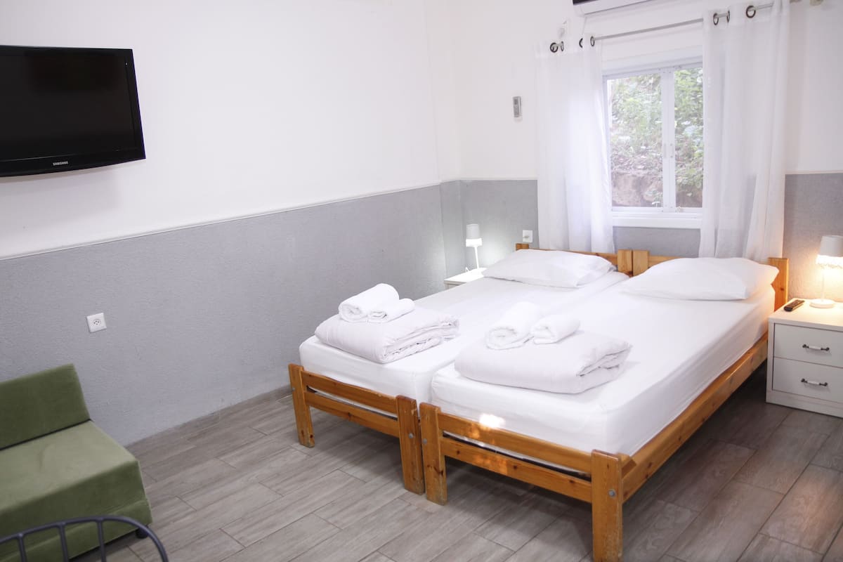 Una cama doble con sábanas blancas, una mesita de noche con lámpara, una silla verde y un televisor montado en la pared.