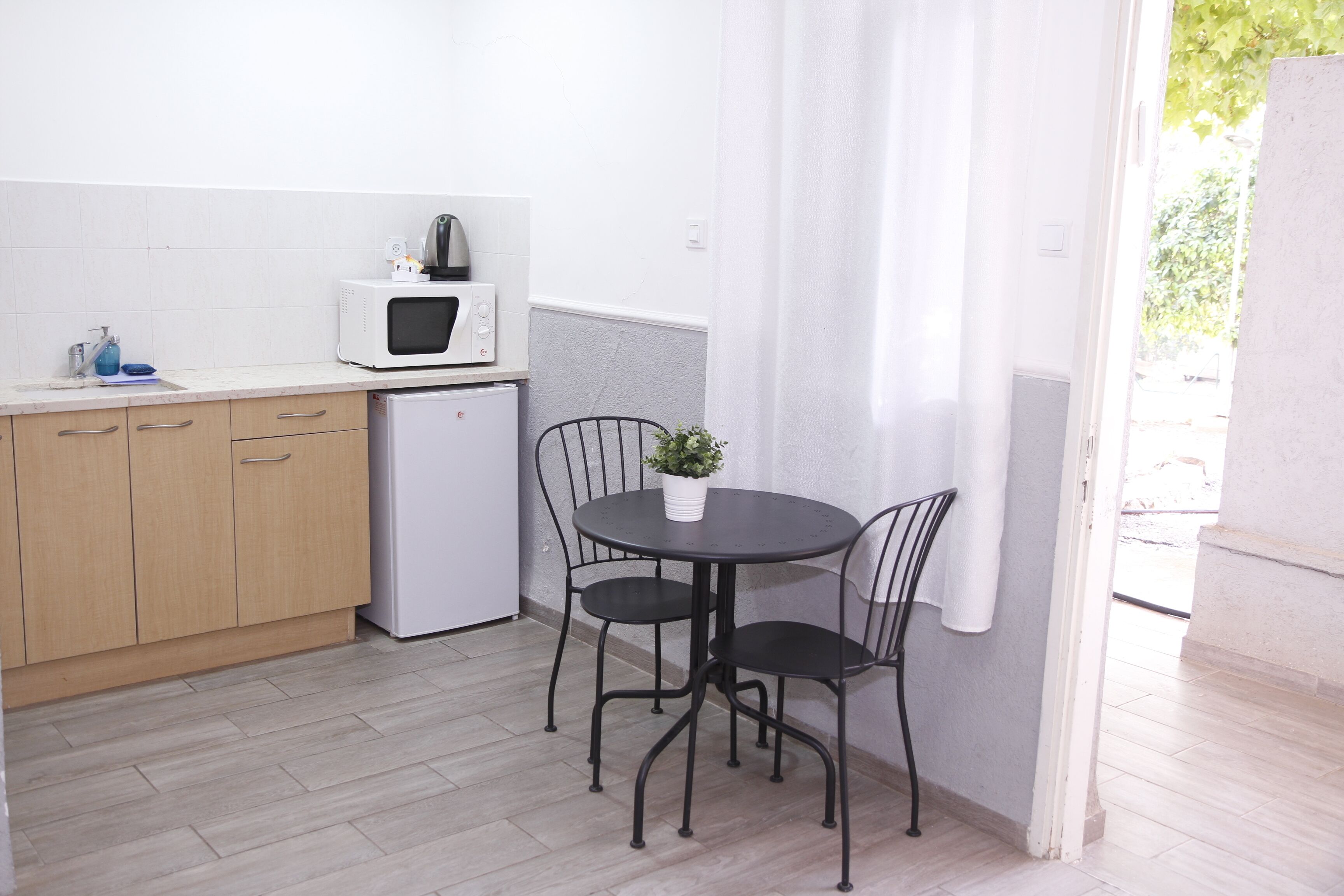 A compact kitchen with a microwave, a small refrigerator, wooden cabinets, a dining table with two chairs, and a potted plant.
