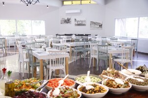 Uma ampla área de jantar com várias mesas e cadeiras dispostas em fileiras. Há diversos recipientes com comida sobre as mesas, e o espaço possui grandes janelas que permitem a entrada de luz natural.