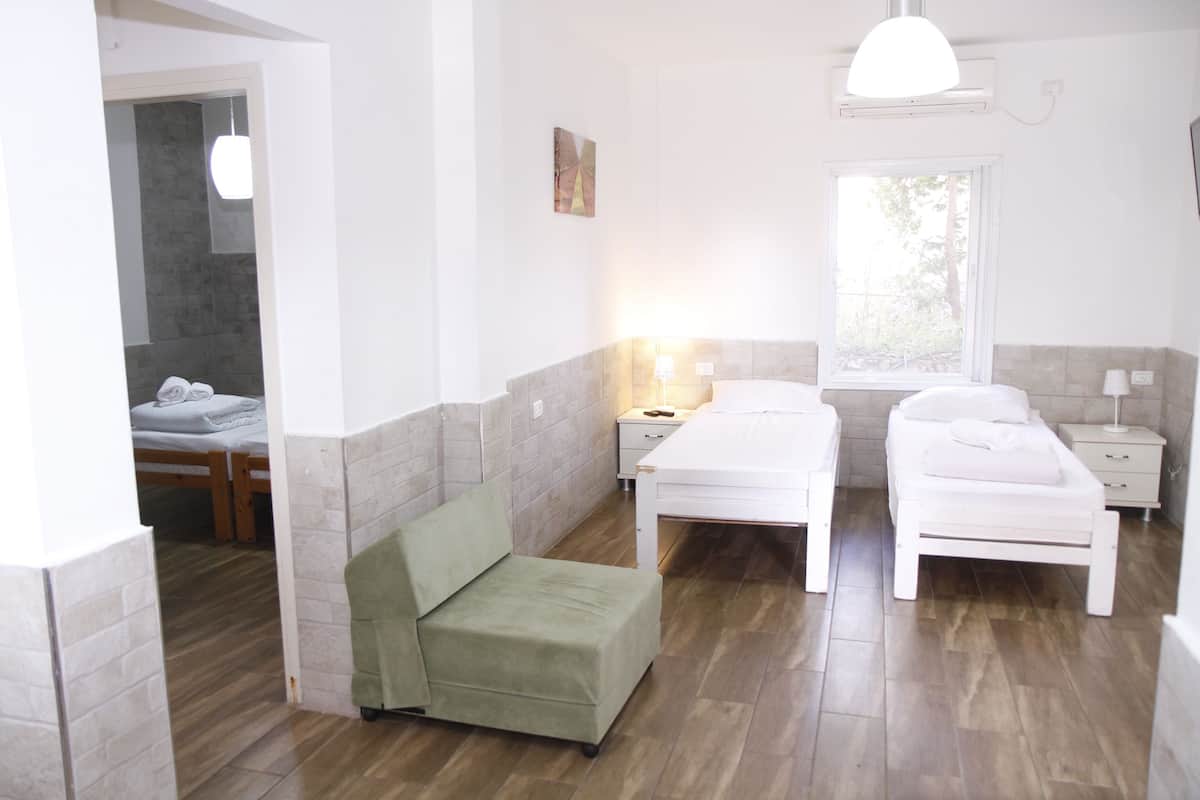 Una habitación con dos camas, una silla verde, una mesita de noche con una lámpara y una ventana.