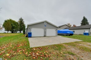 A building with two garage doors, a covered boat, and a grassy area with fallen leaves. - Waterfront Escape on Houghton Lake: Fish & Hike! (Houghton Lake)