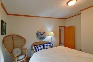 A bedroom with a bed, a wicker chair, a lamp, and a door. - Waterfront Escape on Houghton Lake: Fish & Hike! (Houghton Lake)