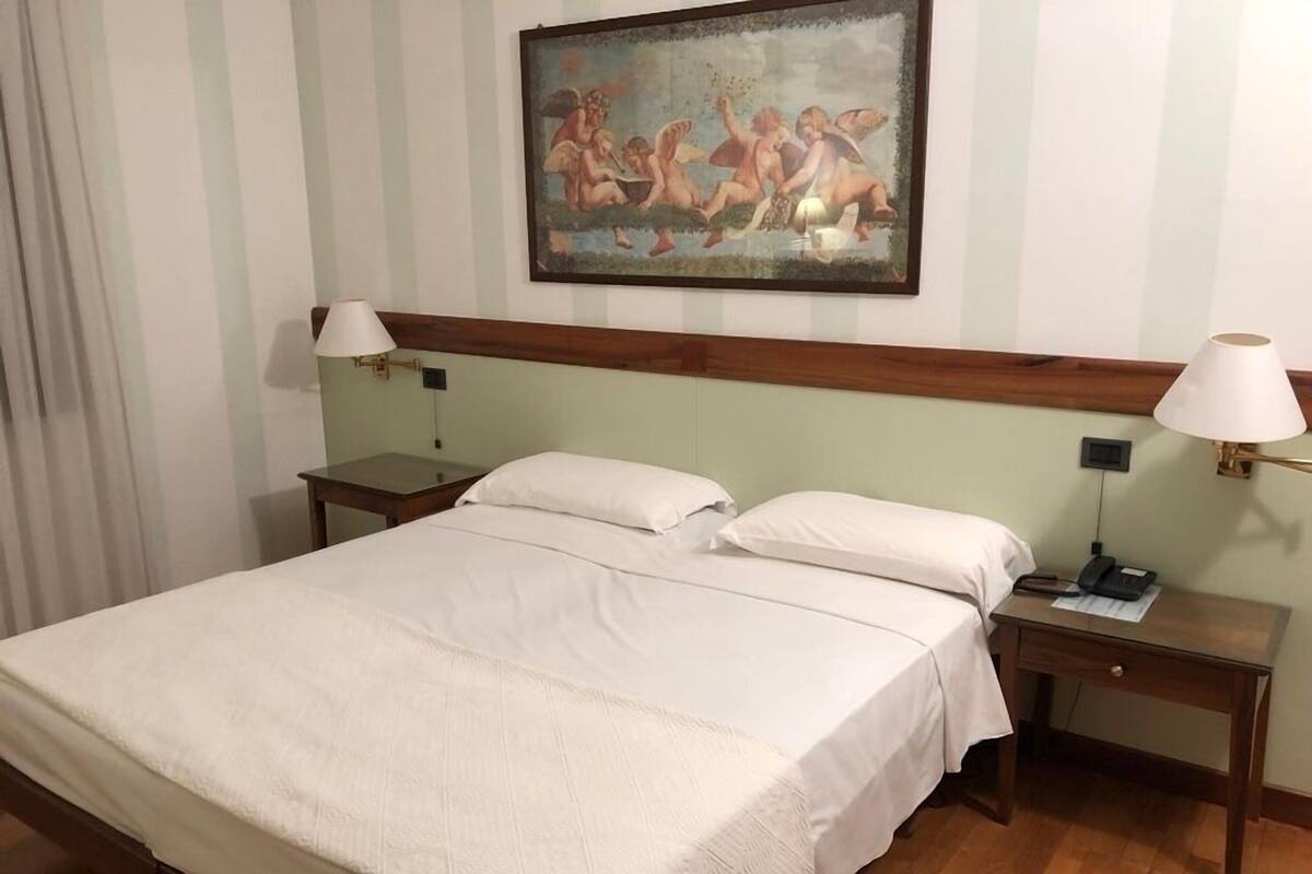 A hotel room with a large bed, two bedside tables, a nightstand, and a painting on the wall.