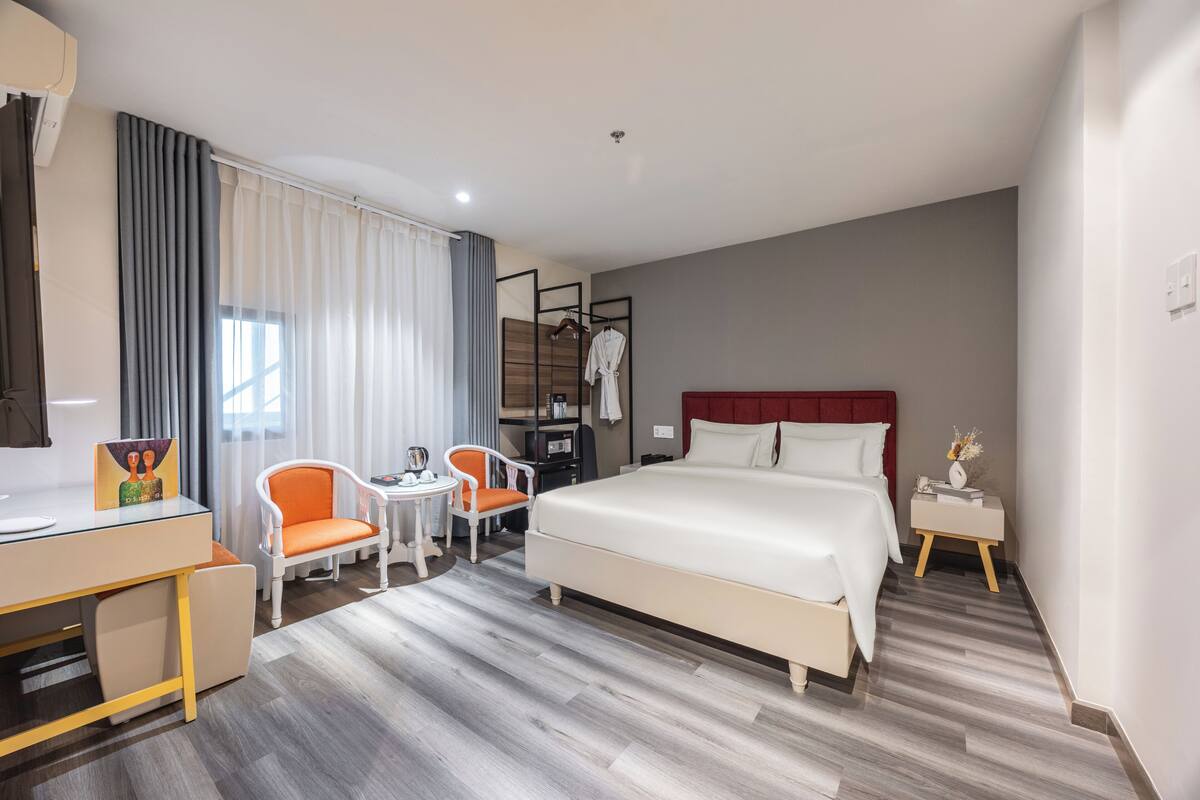 A hotel room with a bed, a desk, two chairs, a small table, a robe hanging, and a nightstand.