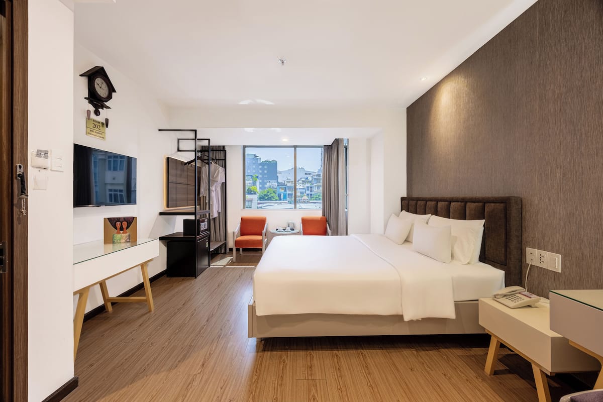 A modern hotel room with a large bed, a desk, a chair, and a view of the city through large windows.