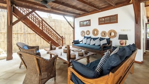 A wooden-floored living area with wicker furniture, a sofa with patterned cushions, and a wall adorned with circular mirrors and a lamp. - The Gili Beach Resort, 4 Bedrooms Villa Gili Trawangan  - Beach Front (Gili Trawangan)