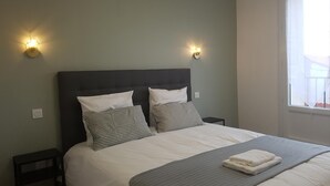 A neatly made bed with white and striped pillows, a grey blanket, and a folded towel on the footboard. There are two bedside tables with lamps, and a window with a view of buildings. - High-end, air-conditioned apartment in the heart of Banyuls (Banyuls-sur-Mer)