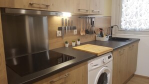 A modern kitchen with a stainless steel range hood, wooden cabinets, a built-in dishwasher, and a window with patterned curtains. - High-end, air-conditioned apartment in the heart of Banyuls (Banyuls-sur-Mer)
