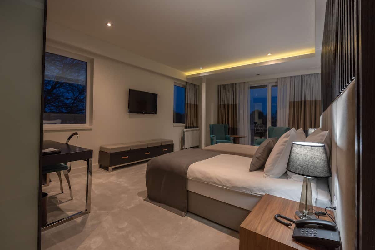 A modern hotel room with a large bed, a desk with a chair, a TV, and a bedside table with a lamp and phone.