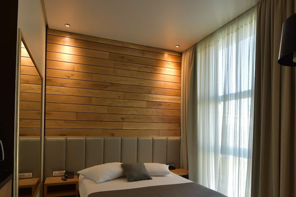 A modern hotel room with a large bed, wooden panel wall, and a large window with curtains.