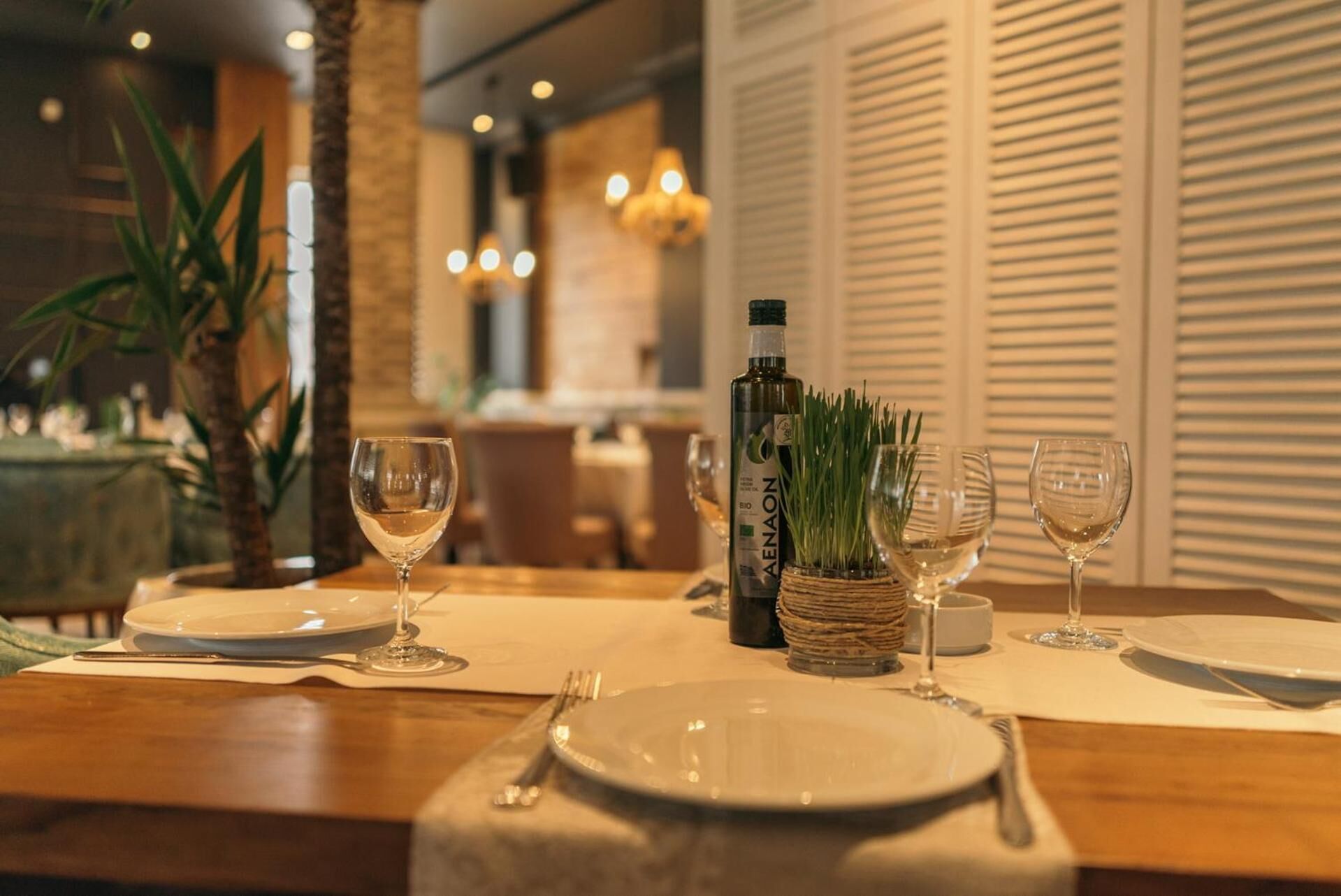 Una mesa de comedor dispuesta con platos, cubiertos y copas de vino, con una botella de vino AENAON y una pequeña planta como centro.