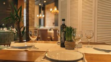 Una mesa de comedor dispuesta con platos, cubiertos y copas de vino, con una botella de vino AENAON y una pequeña planta como centro.