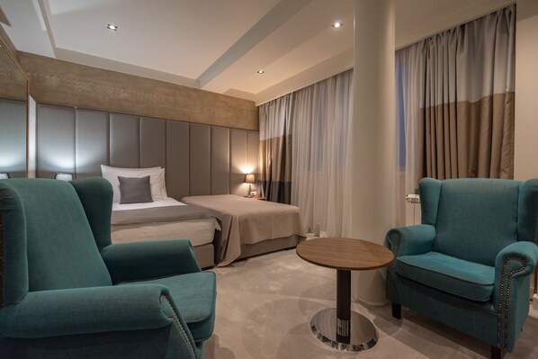 Une chambre d’hôtel moderne avec un lit, deux fauteuils turquoise, une table ronde en bois et une grande fenêtre avec des rideaux.