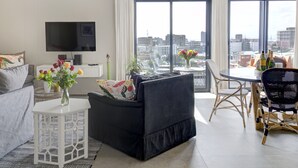 A modern living room with a black sofa, white side table, and a flat-screen TV. There is a dining area with a table and chairs, and a large window offering a city view. - Emily By Stay Amazing (Cape Town)