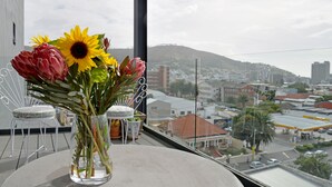 A balcony with a table holding a vase of flowers, a chair, and a view of a cityscape with buildings and a hill. - Emily By Stay Amazing (Cape Town)