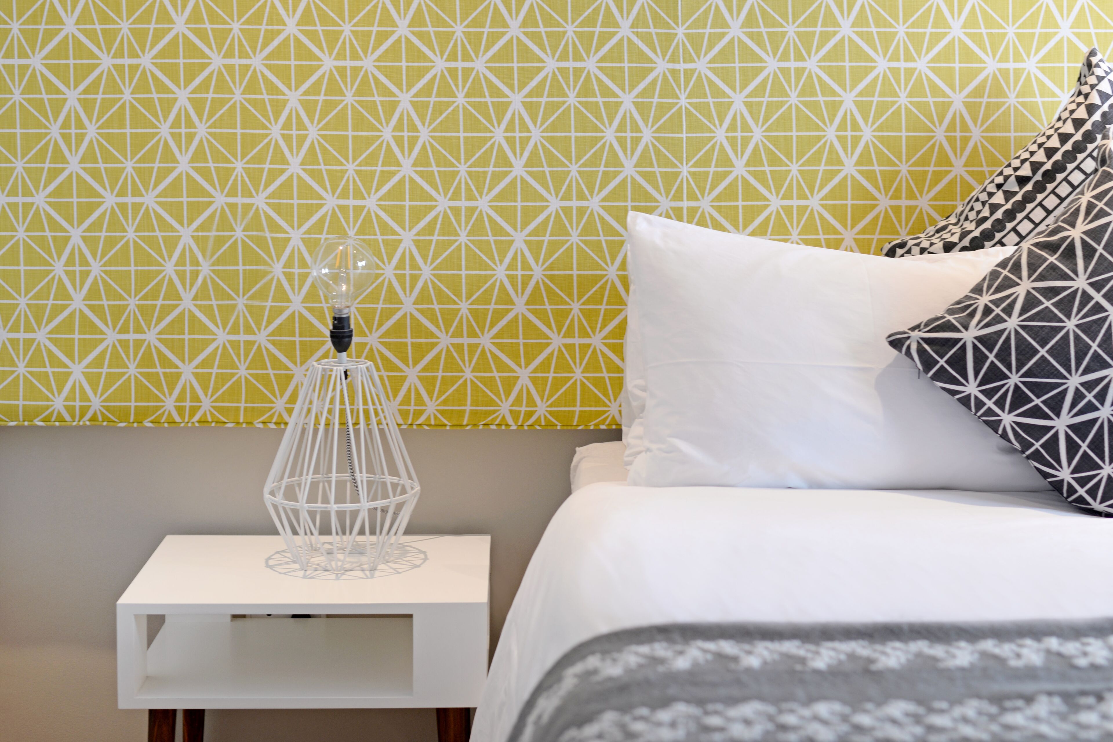 Una cama bien hecha con almohadas blancas y estampadas, una mesita de noche blanca y un papel tapiz geométrico al fondo.