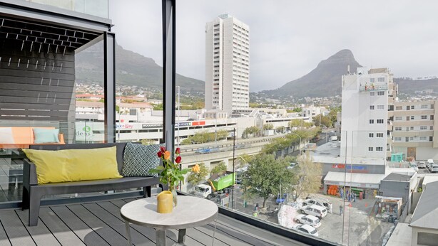 A balcony with a sofa, a small table, and a vase with flowers. A cityscape with buildings and mountains is visible in the background. - Emily By Stay Amazing (Cape Town)