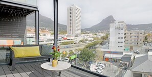 A balcony with a sofa, a small table, and a vase with flowers. A cityscape with buildings and mountains is visible in the background. - Emily By Stay Amazing (Cape Town)