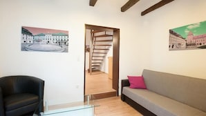 Een moderne woonkamer met een glazen bijzettafeltje, een zwarte fauteuil, een grijze bank met een roze kussen, en twee schilderijen van gebouwen aan de wanden.