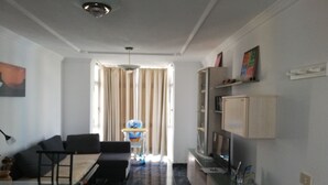 Een moderne woonkamer met een bank, eettafel en stoelen. Er is een keukenhoek met kastjes en een koelkast. Achterin is een raam met gordijnen te zien.