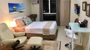 A hotel room with a bed, a desk with a sink, a chair, a lamp, a painting of a beach, and an air conditioning unit. - 27th Flr Condo1 w/WIFI @ IT Park Avida Riala Twrs (Cebu City)