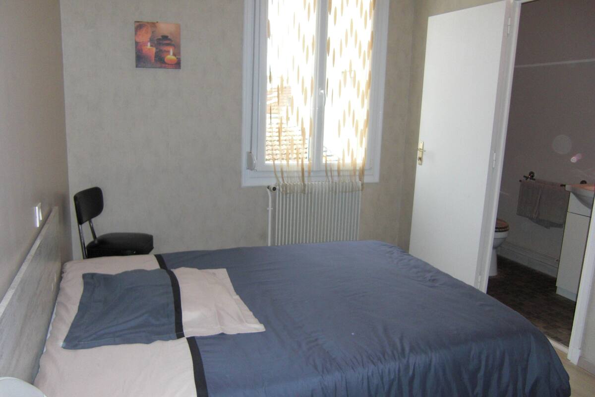 A bedroom with a bed, a chair, a radiator, a window with blinds, and a door leading to a bathroom.
